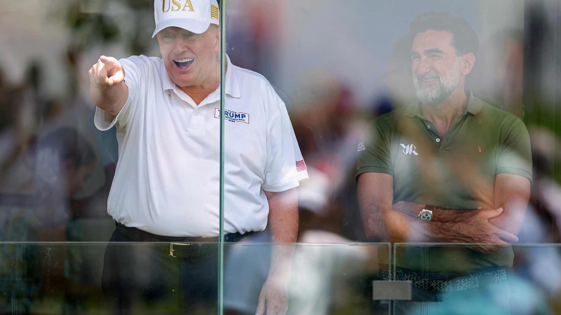 Two men stand behind glass; one in a white USA hat and golf attire is pointing and smiling, while the other in a green shirt stands with arms crossed, both facing the same direction.