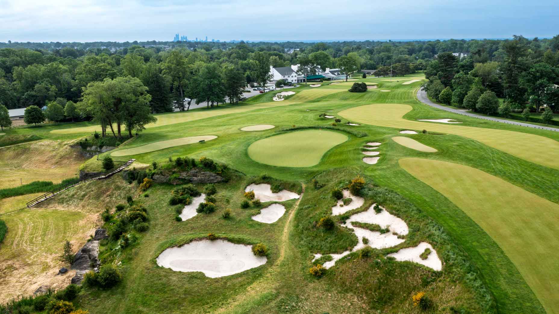 Aerial view of Merion golf course with sand bunkers, green fairways, and trees bathed in the gentle glow of the sun. Clubhouse buildings are visible in the background, with a city skyline on the horizon under a cloudy sky.