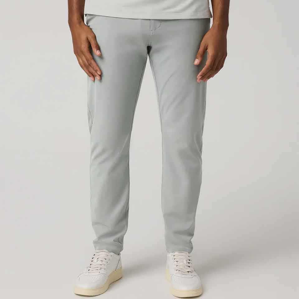A person stands against a plain background, wearing light gray Vuori slim-fit pants and white sneakers, with hands relaxed at their sides. The upper body is mostly out of frame.