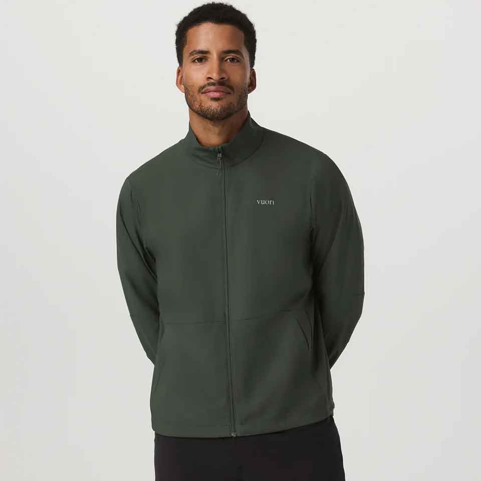 A man with short hair and a beard wears a dark green Vuori zip-up jacket, standing against a plain white background with his hands behind his back.