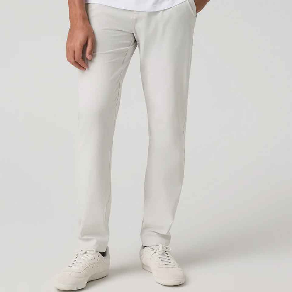 A person wearing Vuori white pants and white sneakers stands with one hand in their pocket against a plain light background. Their upper body is mostly out of frame, making it a subtle nod to golf apparel perfect for Father's Day.