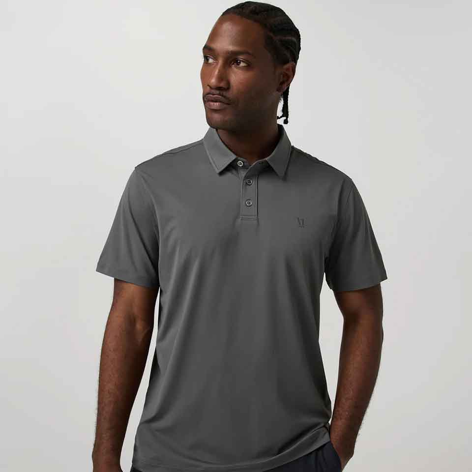 A man with braided hair wears a short-sleeve, gray Vuori polo shirt&mdash;perfect golf apparel for Father&rsquo;s Day&mdash;as he stands against a plain, light background, looking slightly to his left with one hand in his pocket.