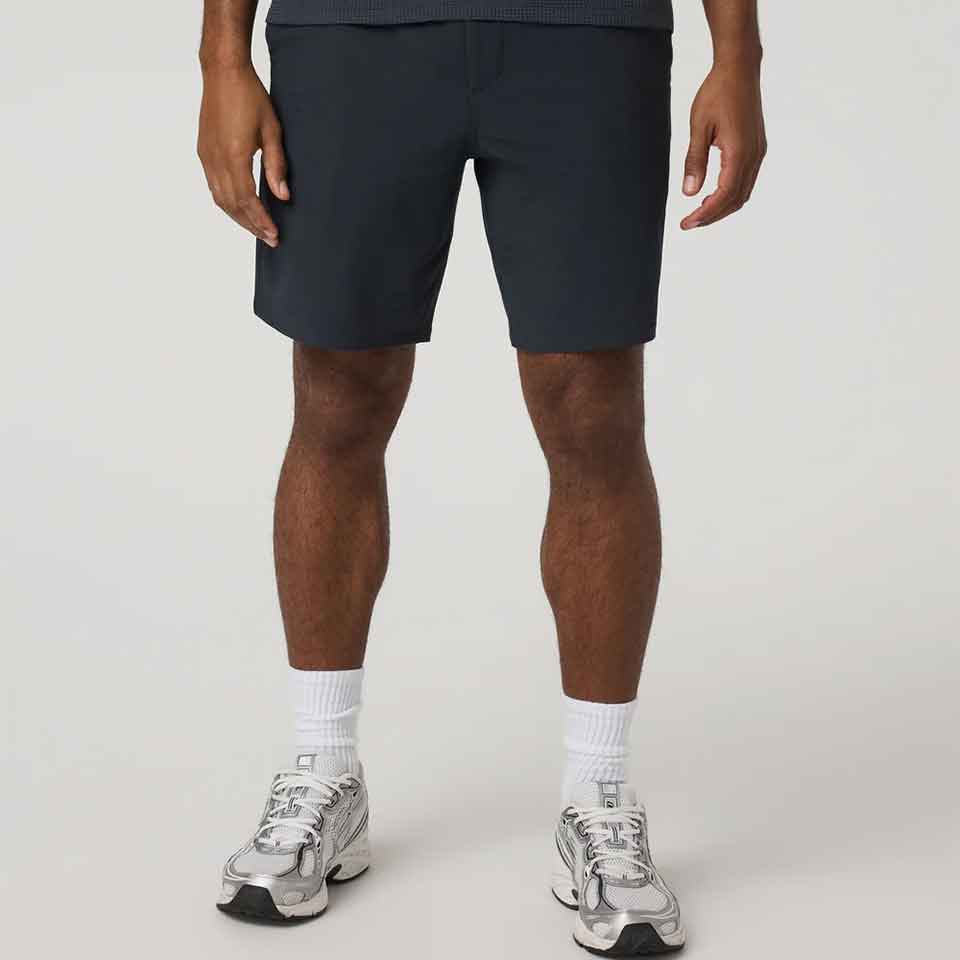 A person stands against a plain background, wearing Vuori dark shorts, white crew socks, and silver athletic sneakers&mdash;their upper body is not visible in the image.