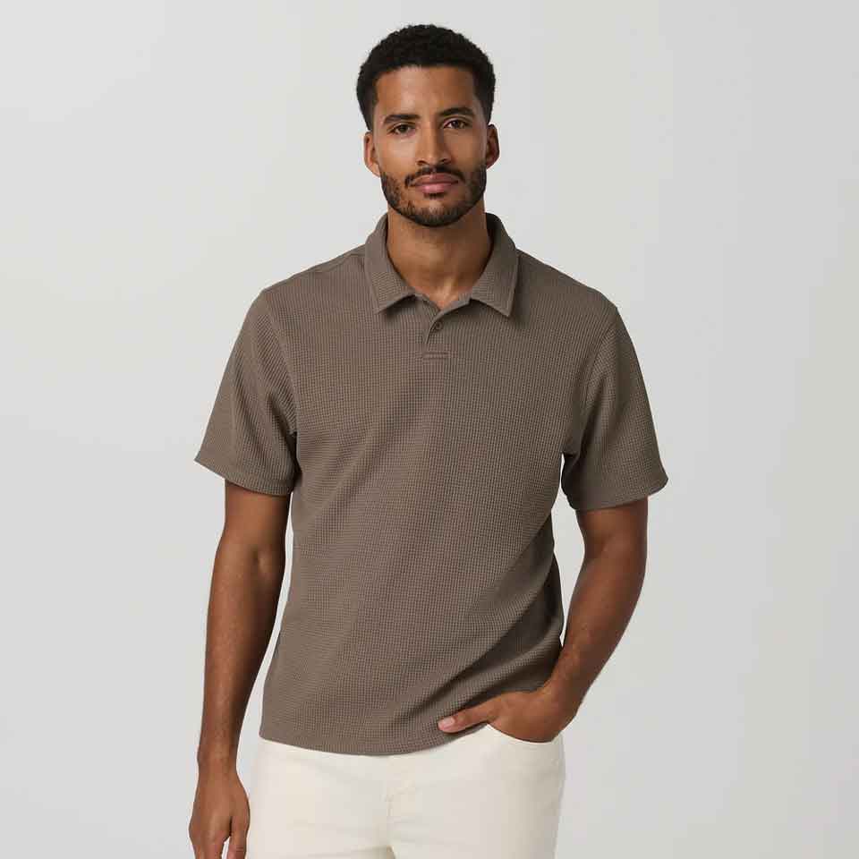A man with short hair and a beard wears a textured brown Vuori polo shirt and light-colored pants, standing against a plain light gray background. He has one hand in his pocket and a neutral expression&mdash;perfect Father's Day golf apparel.