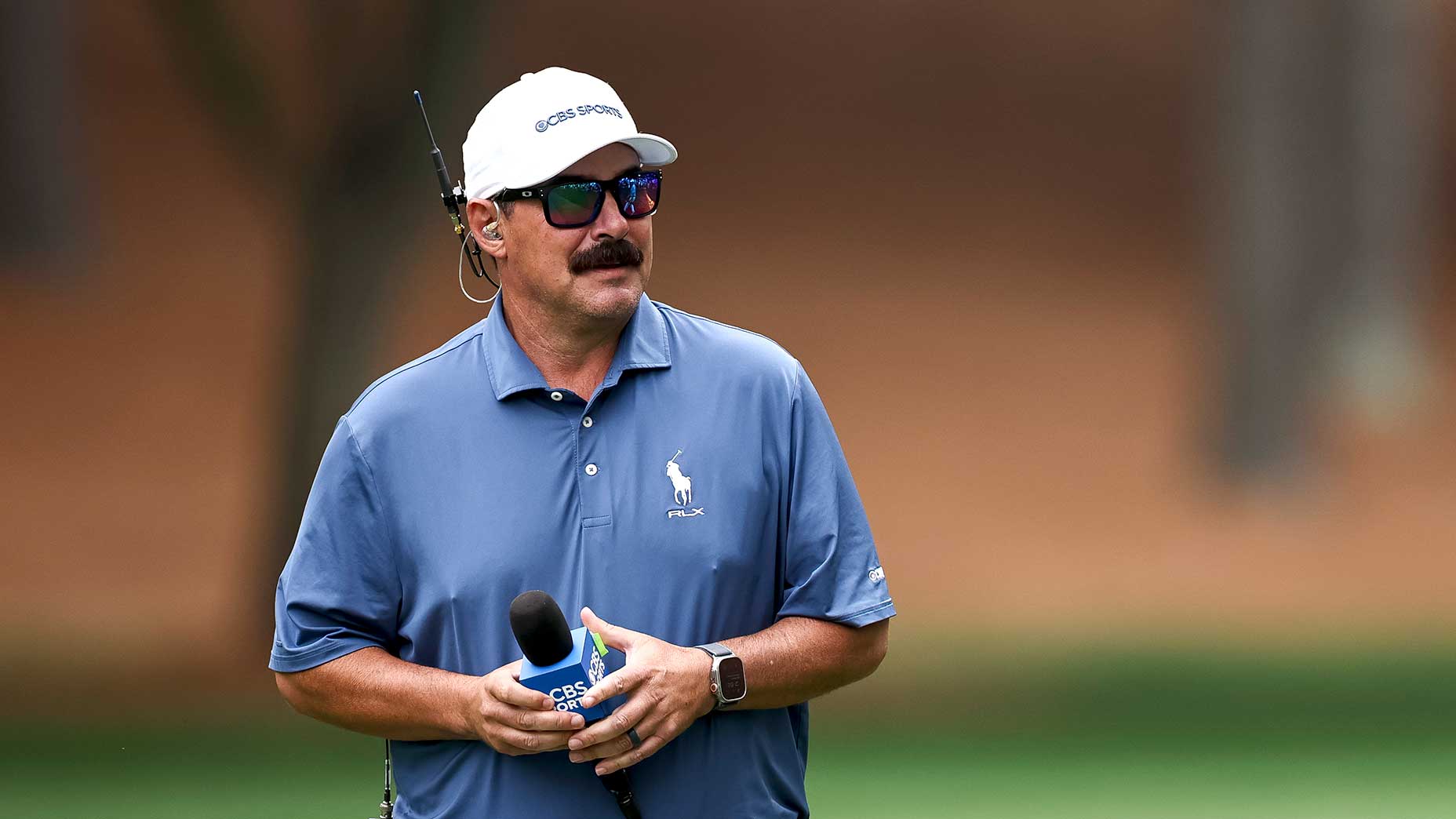Johnson Wagner, wearing sunglasses, a headset, and a blue polo shirt, holds a CBS Sports microphone while standing outdoors on a golf course.