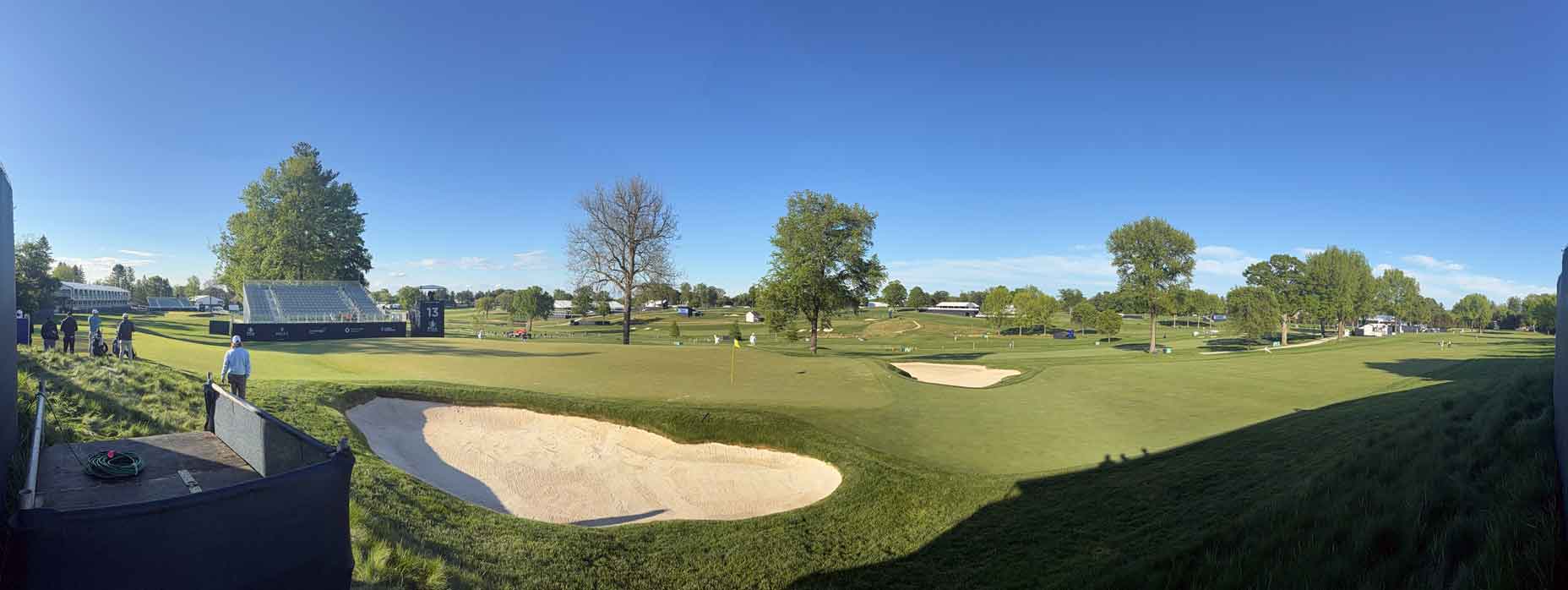 A panoramic view of Aronimink golf course under a clear blue sky, featuring green fairways, sand bunkers, scattered trees, and a few people and structures in the distance&mdash;ideal for early observations ahead of the PGA Championship.