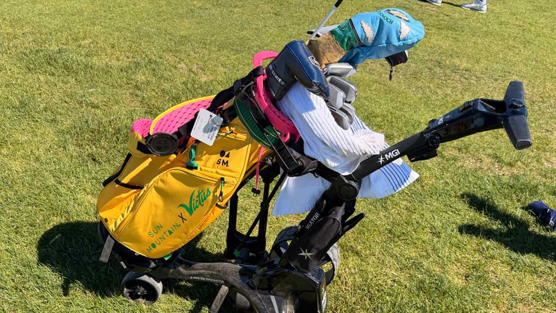 A yellow Sun Mountain golf bag sits on a black pushcart on green grass, filled with clubs and head covers, with a white towel draped over the top. This stylish pencil golf bag is perfect for any round.
