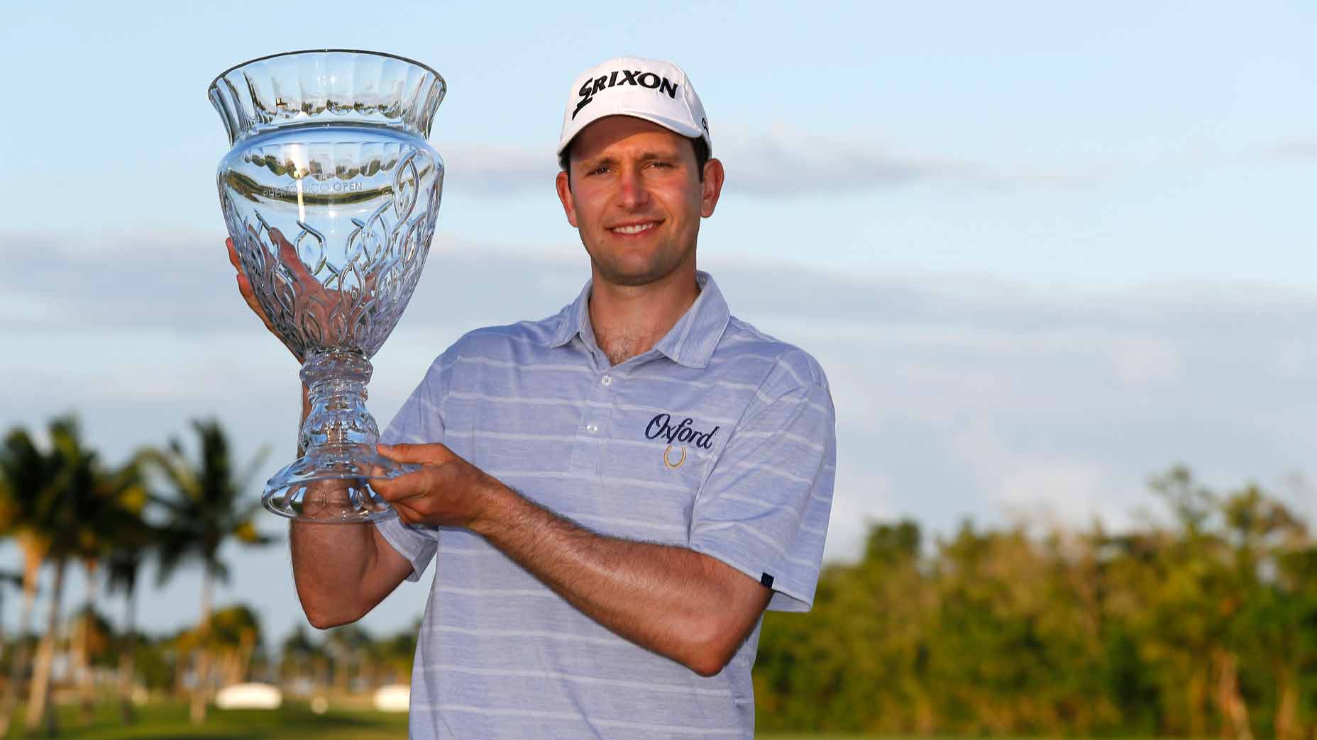 A retiring pro in a striped golf shirt and cap smiles while holding a large crystal trophy outdoors, with palm trees and greenery behind him&mdash;celebrating his career and sharing his golf secrets from years on the PGA Tour.