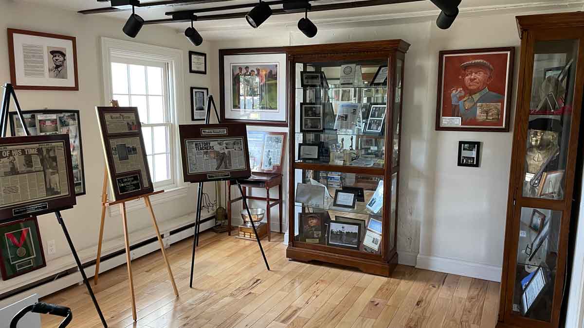 A small museum room with framed photos and newspaper clippings on easels, including a display on a forgotten hero of the U.S. Open, with memorabilia-filled cases and portraits on light wooden floors lit by natural window light.