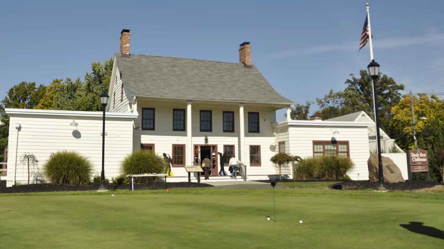 A white colonial-style building with columns, two chimneys, and people entering sits behind a green lawn with a putting area&mdash;a nod to the U.S. Open; an American flag flies on a tall pole to the right.