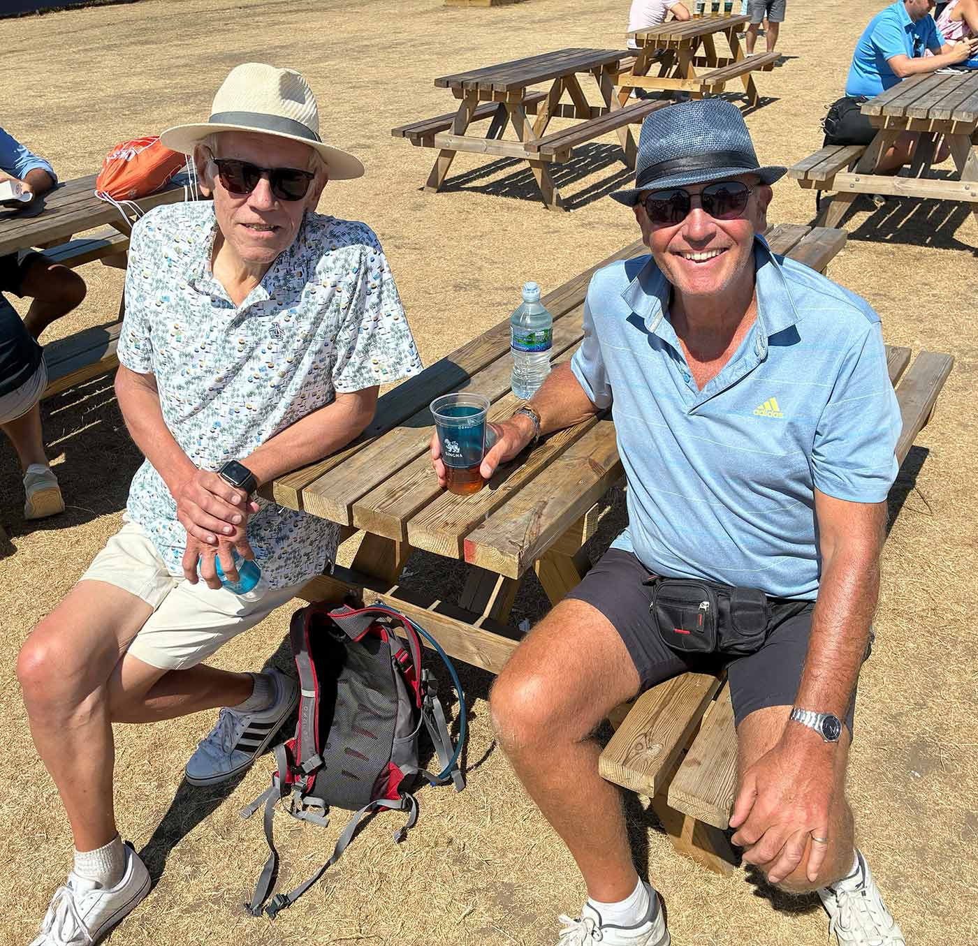 Two men in hats and sunglasses sit on a wooden picnic bench outdoors on a sunny day, holding drinks and smiling. A backpack sits nearby, and open spectators can be seen in the background, adding to the lively atmosphere.