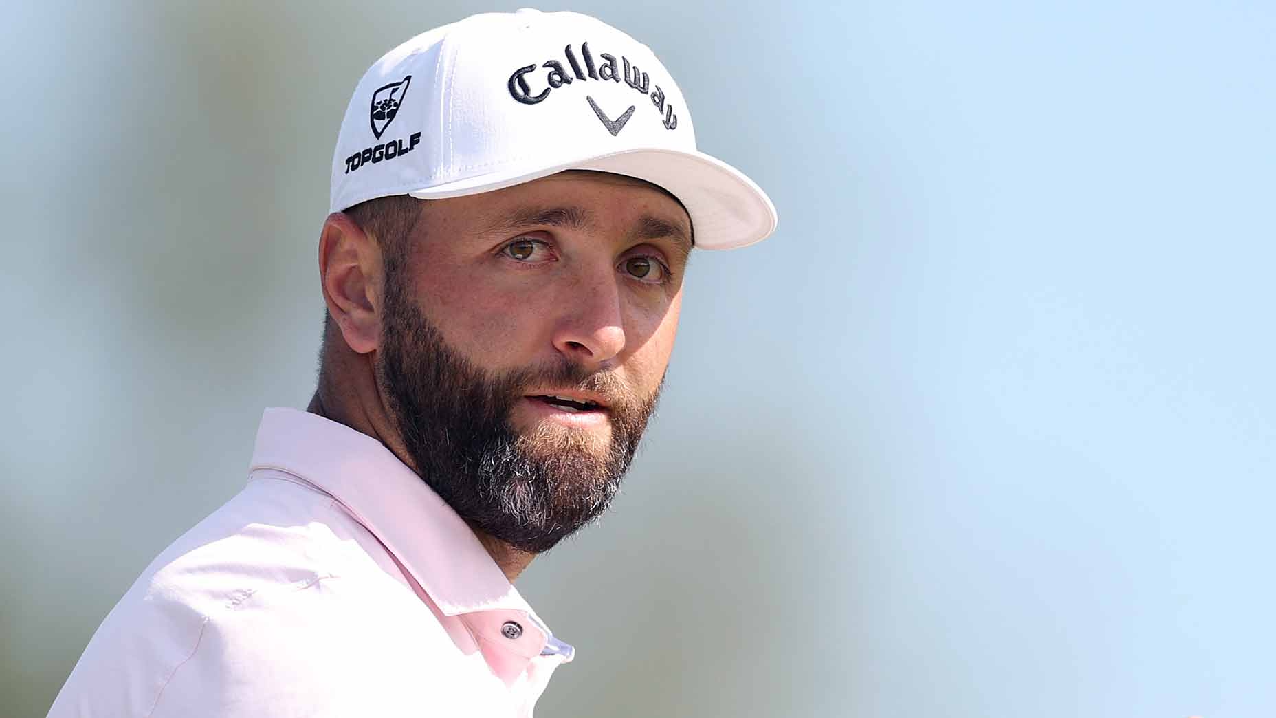 A man with a beard wearing a white Callaway golf cap and a light pink collared shirt looks to the side against a blurred outdoor background.