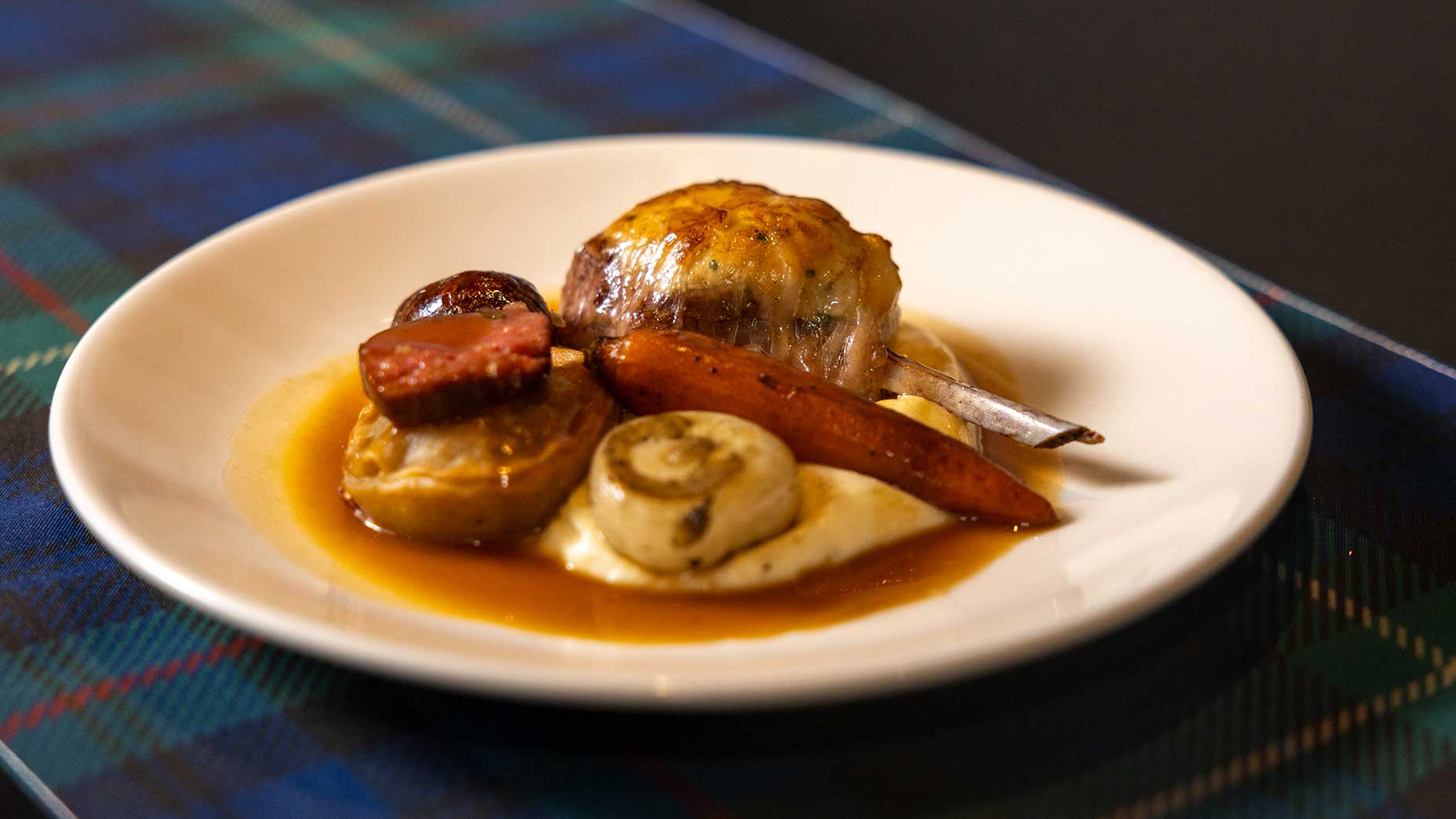 A white plate holds roasted meat, a carrot, mushrooms, a potato, and gravy, all served atop a bed of creamy mashed potatoes&mdash;an inviting showcase of Scottish fare&mdash;set on a blue tartan tablecloth.