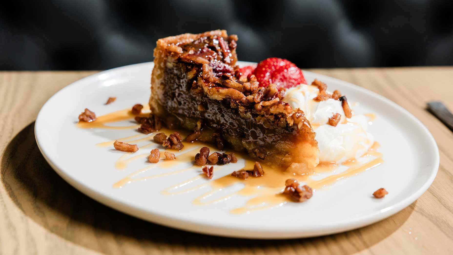 A slice of pecan pie reminiscent of classic Southern and Scottish fare, topped with caramel sauce, chopped pecans, whipped cream, and strawberries, is artfully presented on a white plate set atop a wooden table&mdash;an indulgent treat worthy of McLemore Resort&rsquo;s dessert menu.