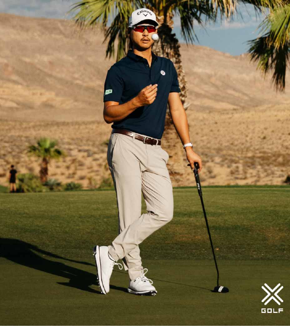 A golfer in a navy shirt and beige pants stands on a green, holding a putter and wearing sleek Payntr shoes. Palm trees and desert mountains are in the background. The X Golf logo is in the corner.