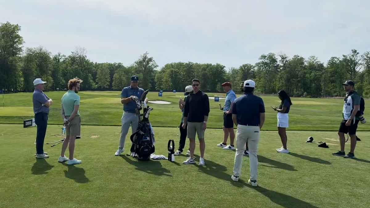A group of people stands on a golf course near golf bags and clubs, talking and preparing to play. Trees and practice areas are visible in the background on a sunny day, marking the first event of a new era in LIV golf.