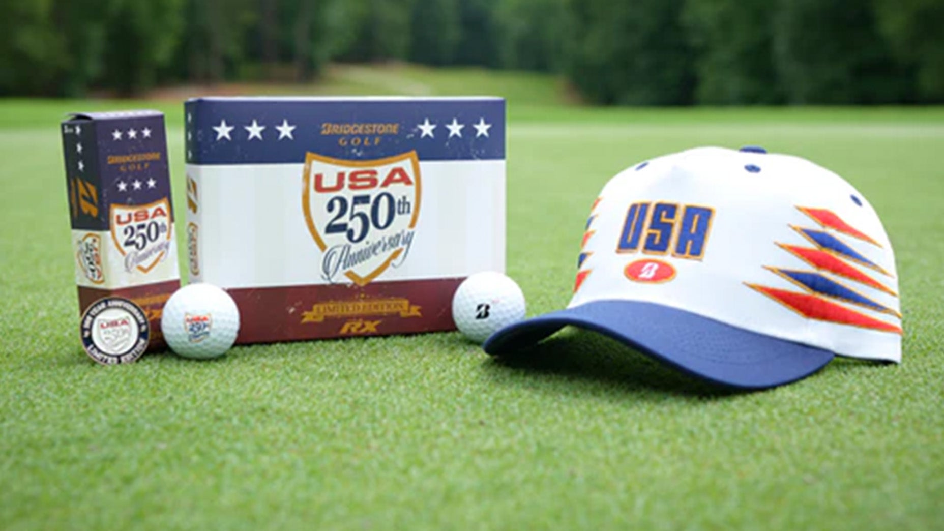 Golf gear on green grass: a USA 250th Anniversary Bridgestone golf ball box, red white and blue gear including two golf balls, a ball marker, and a white cap with USA in blue and orange, with trees blurred in the background.
