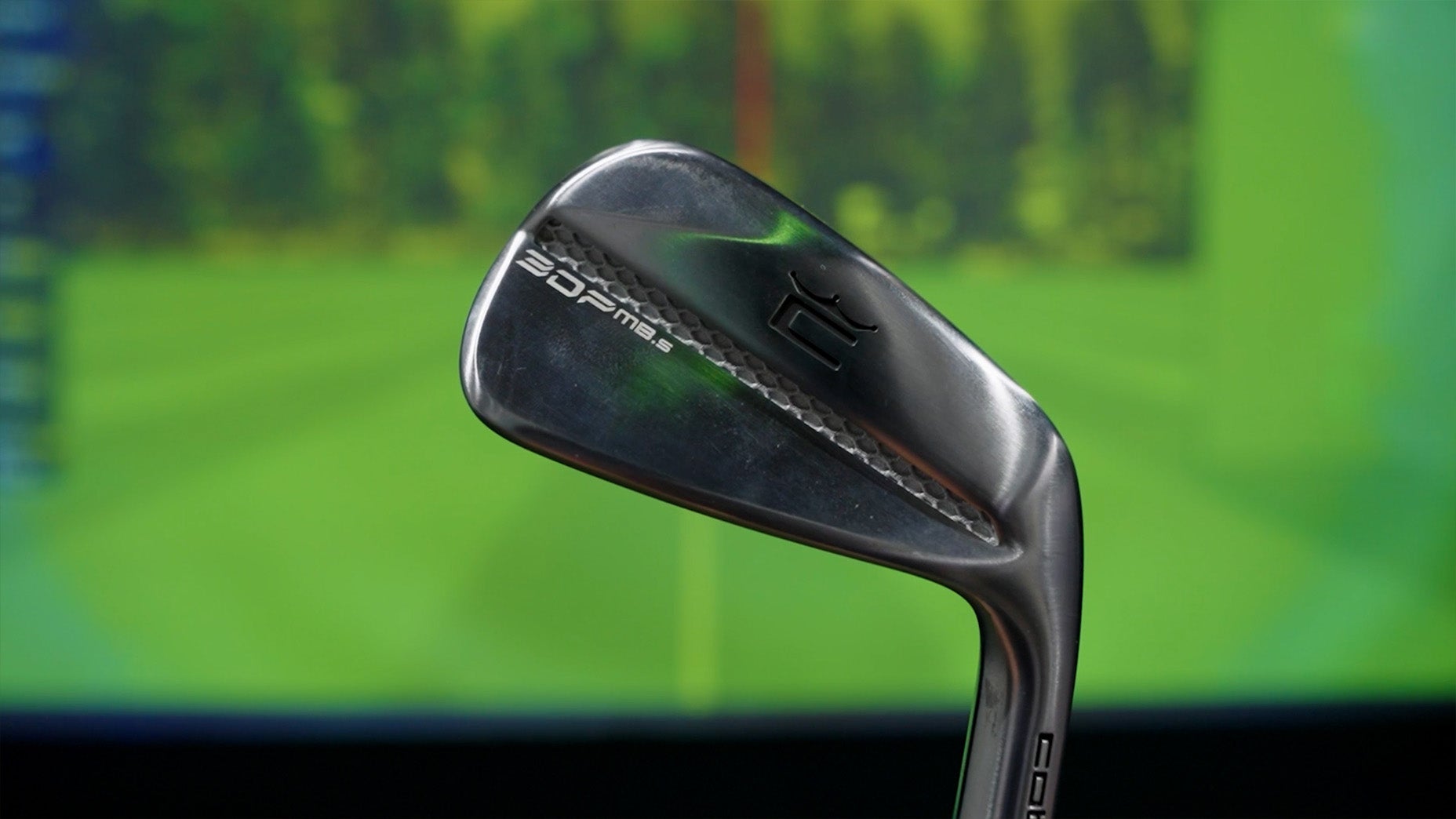 A close-up of a metallic golf iron with the label 2021 MB against a blurred background of a green golf course simulation.