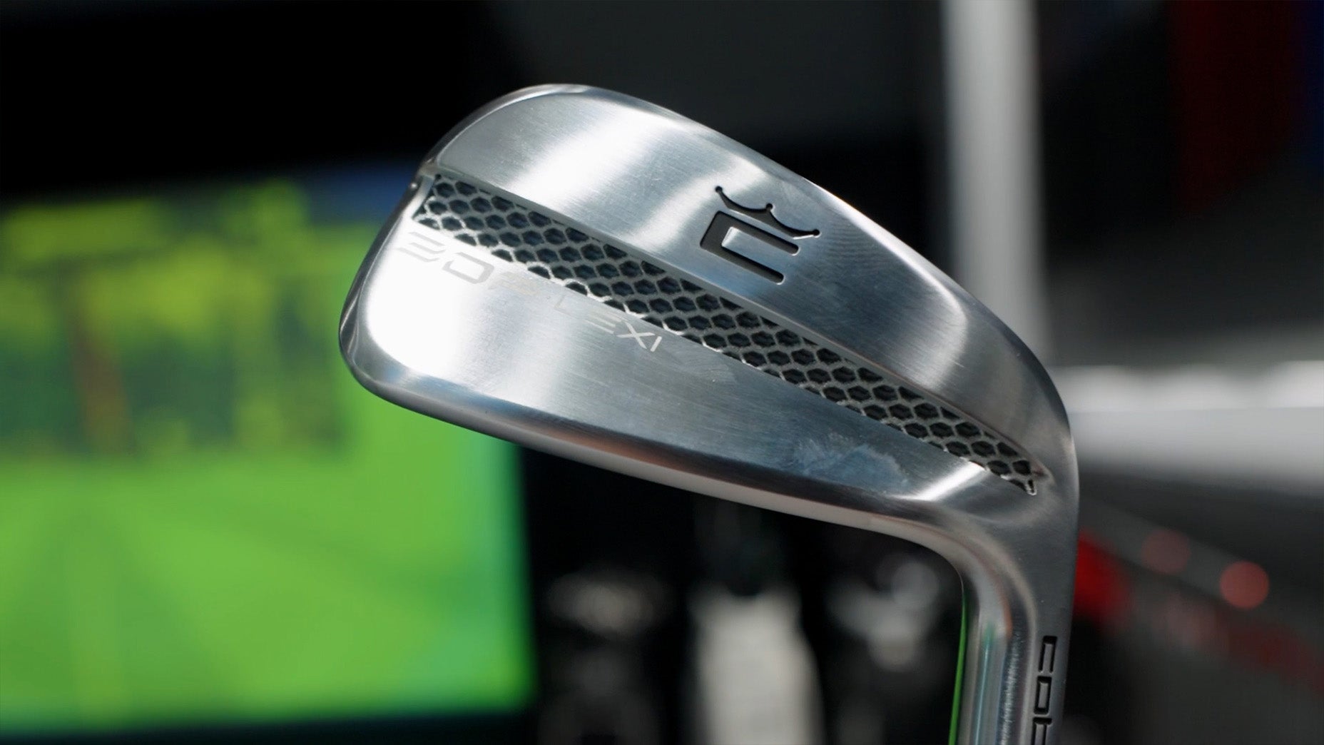 A close-up of a shiny metal golf club iron, featuring a textured honeycomb pattern and engraved branding on the club head, with a blurred green screen in the background.