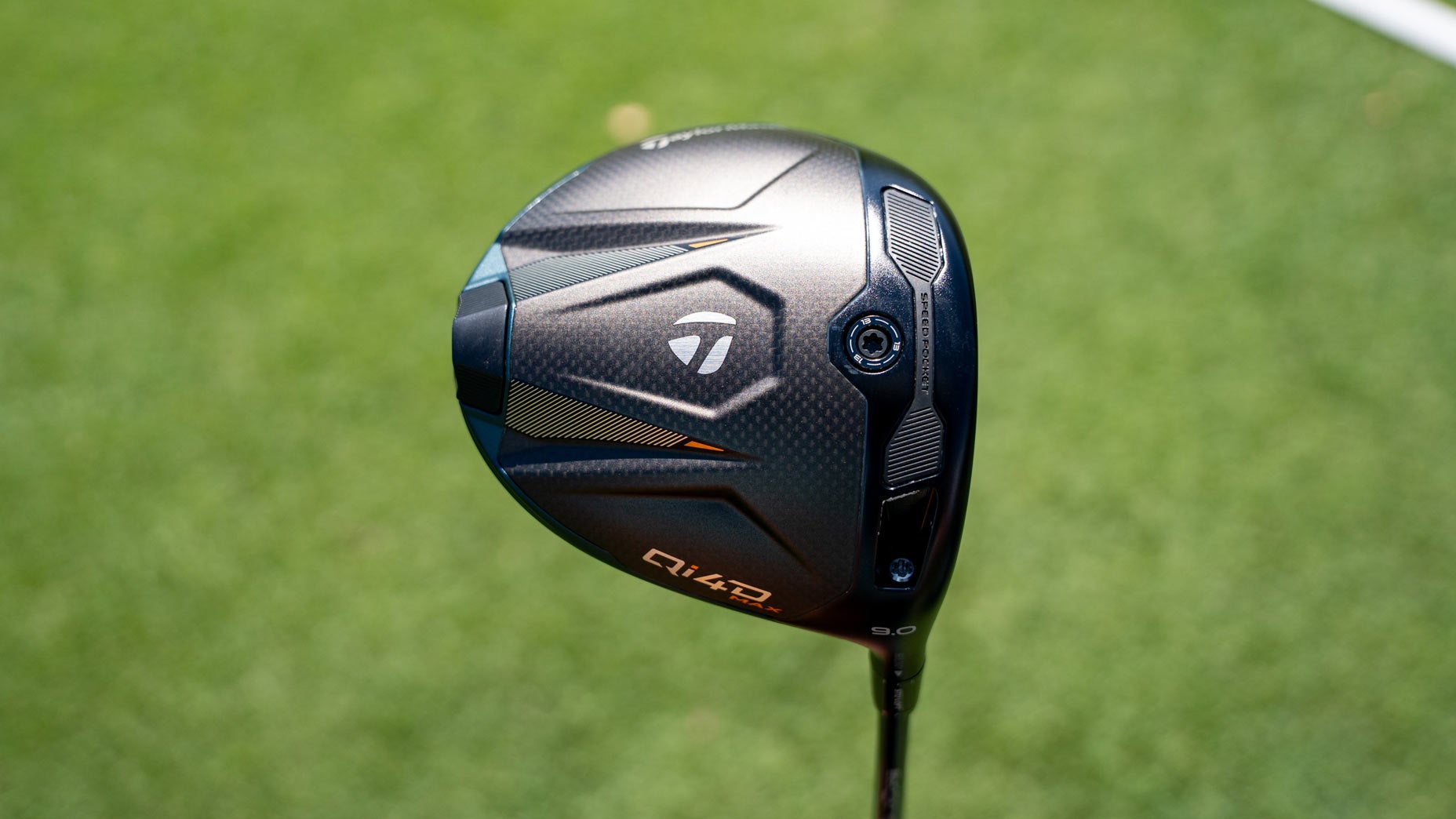 A close-up of a TaylorMade golf driver clubhead with a dark, textured design, silver and orange accents, positioned against a blurred green grass background&mdash;just like you&rsquo;d spot in the latest Tour Report from the PGA Tour.
