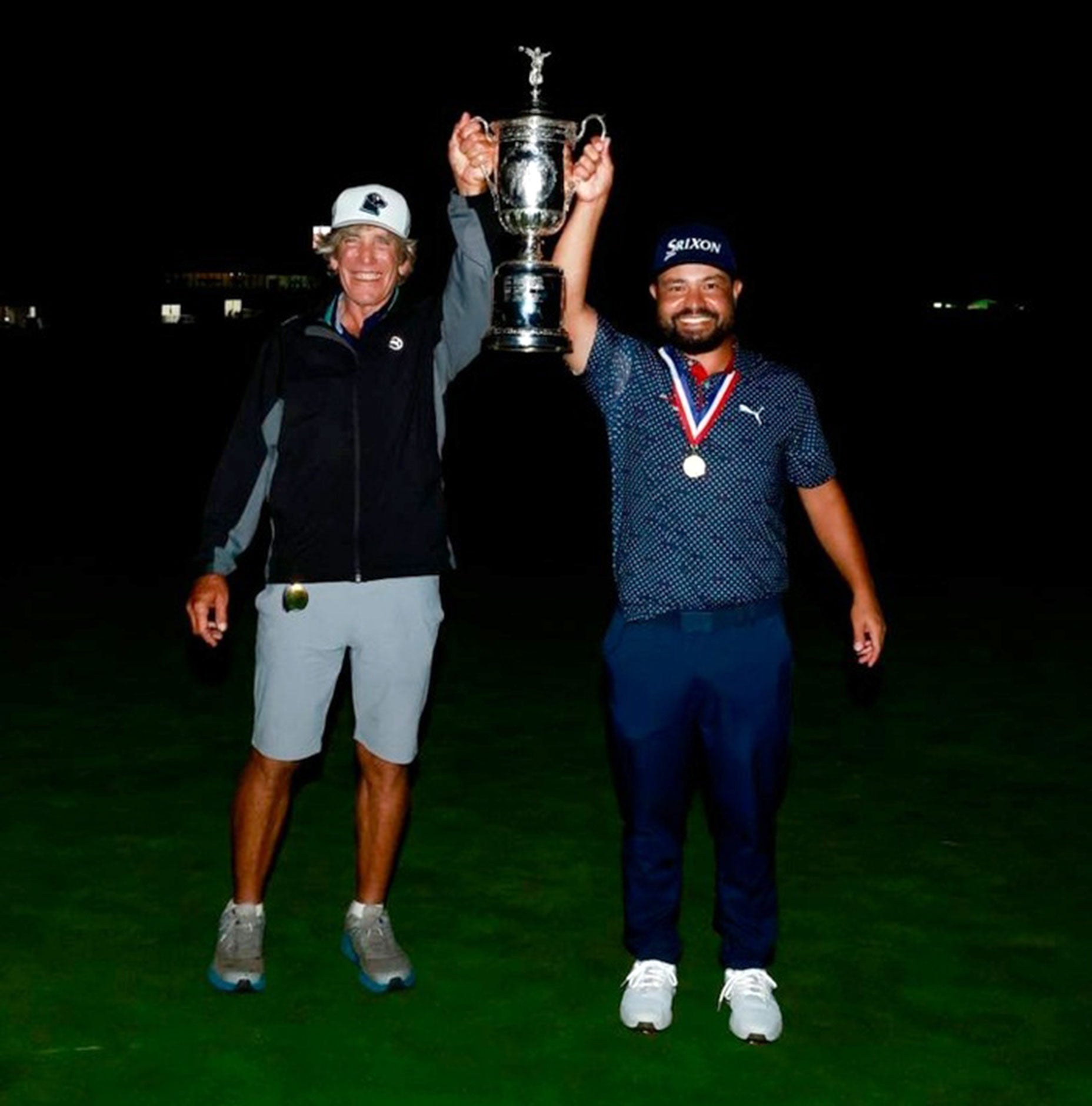 Two men stand together at night on a golf course, smiling and holding up a large silver trophy. One wears a golf hat and vest; the other sports a KTKT cap, medal, and classic golf attire.