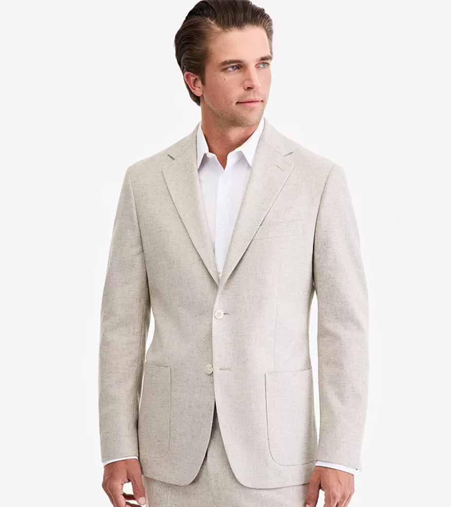 A man with short brown hair is wearing a stylish beige suit jacket and white dress shirt, standing against a plain white background and looking slightly to his left.