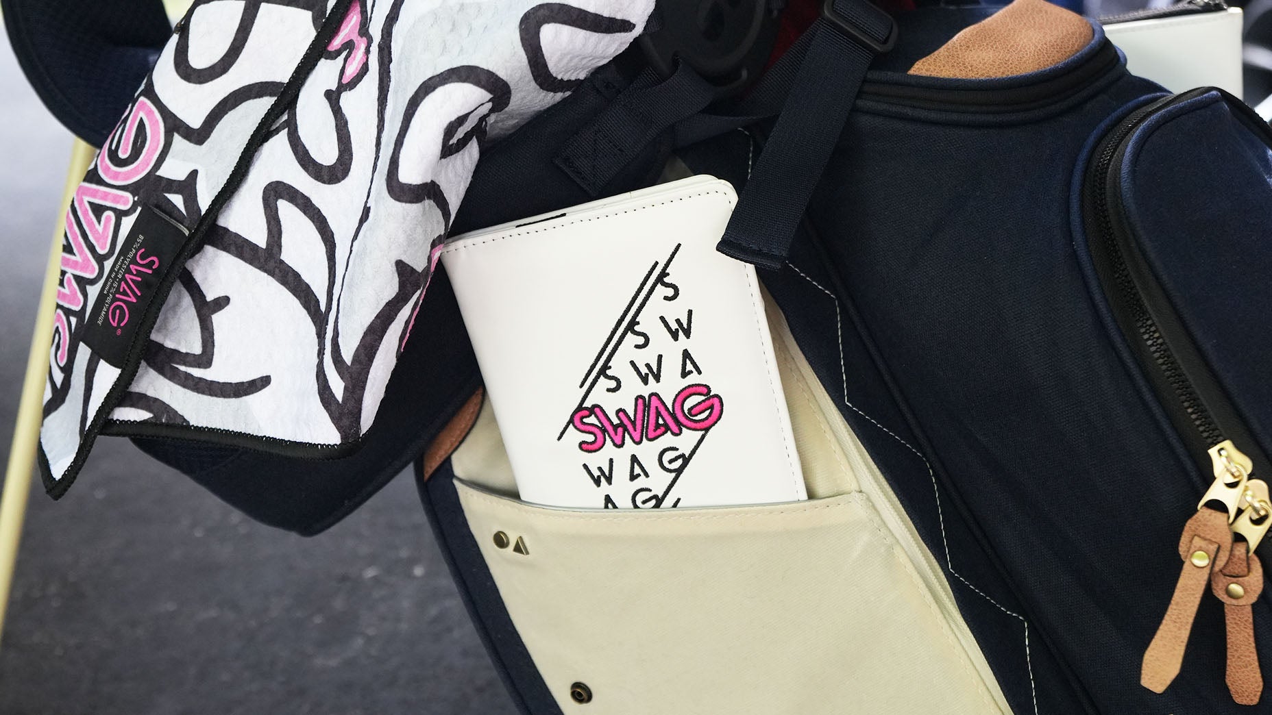 A close-up of a golf bag pocket holding a white cover with SWAG printed in black and pink letters, accompanied by a black and white towel with the same logo draped nearby, showcases the latest SWAG accessories that add style and flair to your game.