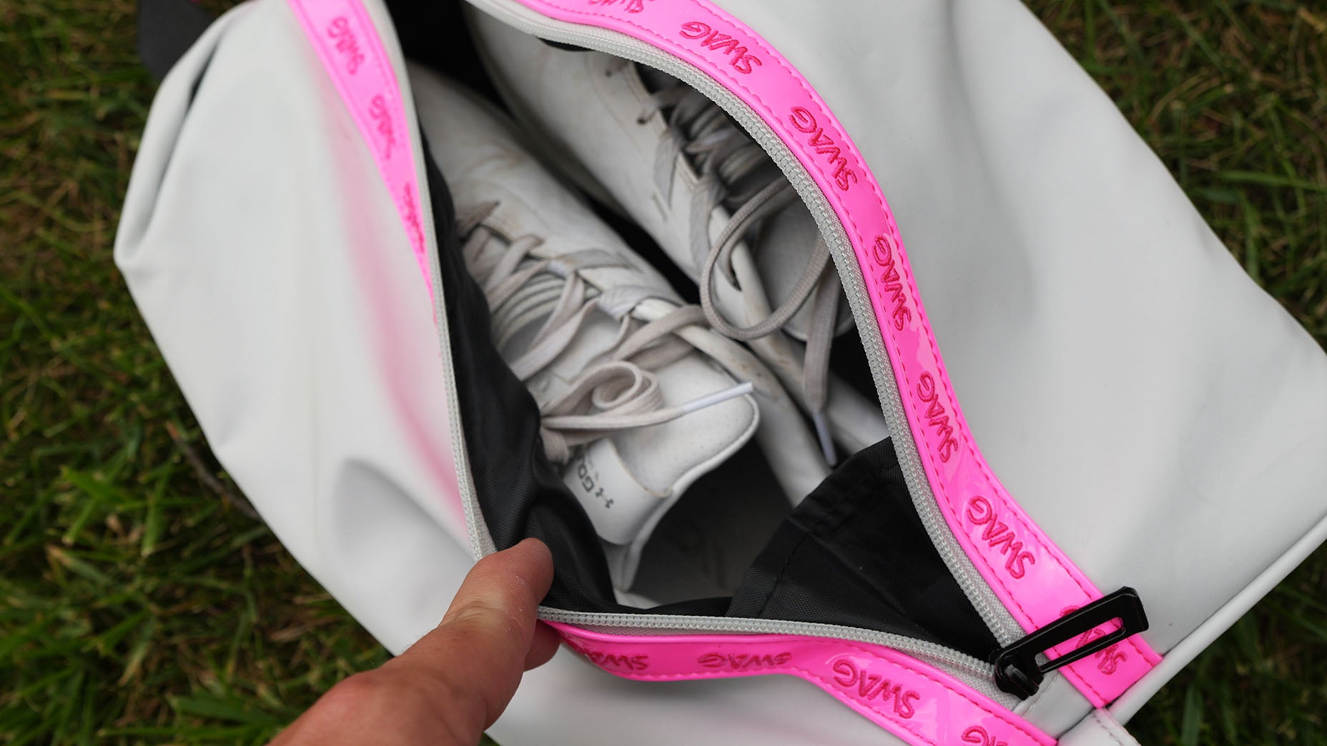 A hand opens a white bag with a pink zipper labeled SWAG, revealing a pair of white sneakers&mdash;stylish SWAG accessories&mdash;inside. The bag rests on green grass.