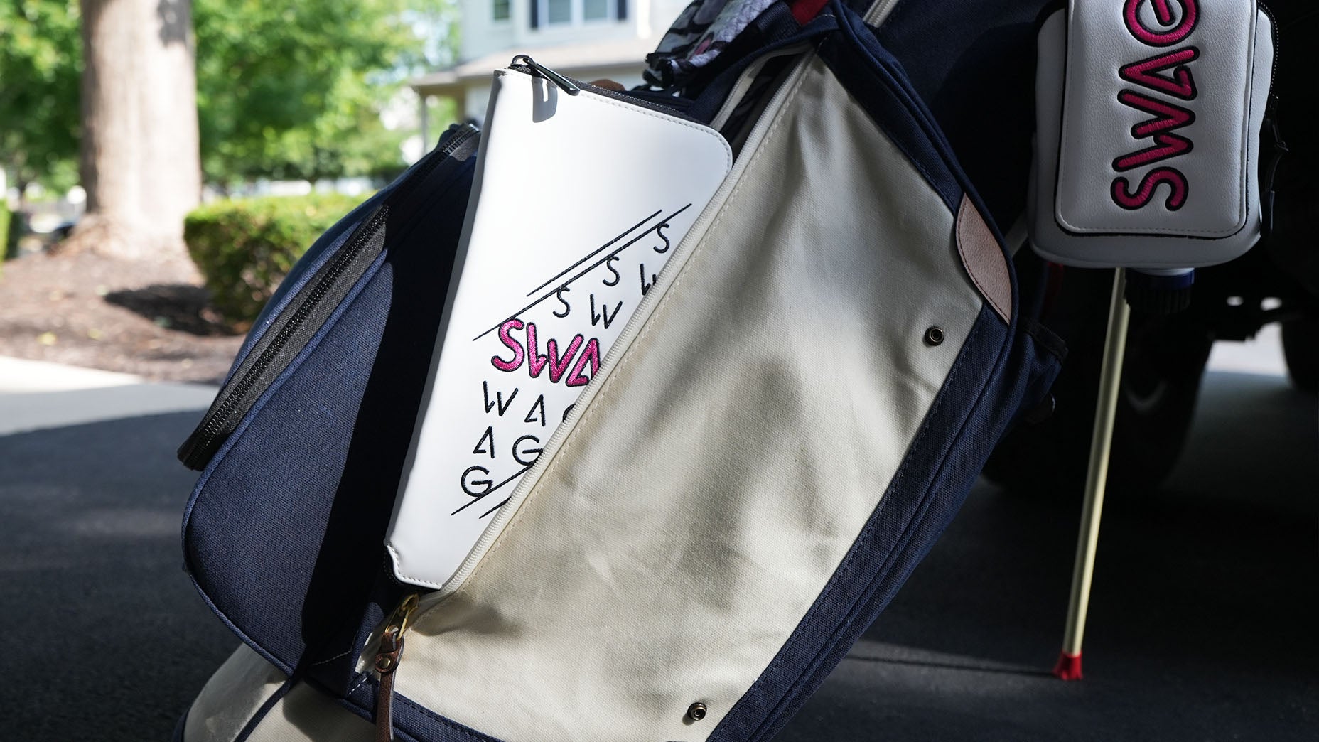 A close-up of a golf bag with a white pouch reading SWAG multiple times in pink and black letters, hanging on the side&mdash;a perfect example of the latest SWAG accessories. The background features trees, bushes, and a house.