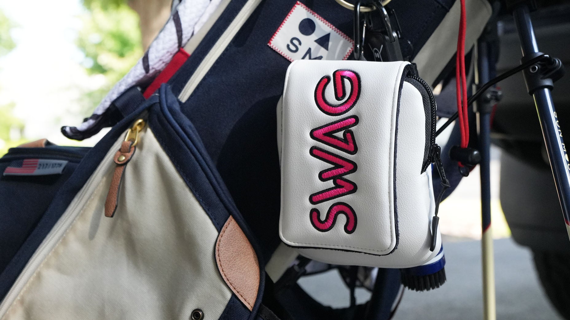 A white pouch with SWAG in bold pink letters&mdash;one of the latest SWAG accessories&mdash;hangs from a navy and beige golf bag. The bag is adorned with various patches, making it a standout among fashion accessories, and the scene is outdoors with greenery in the blurred background.