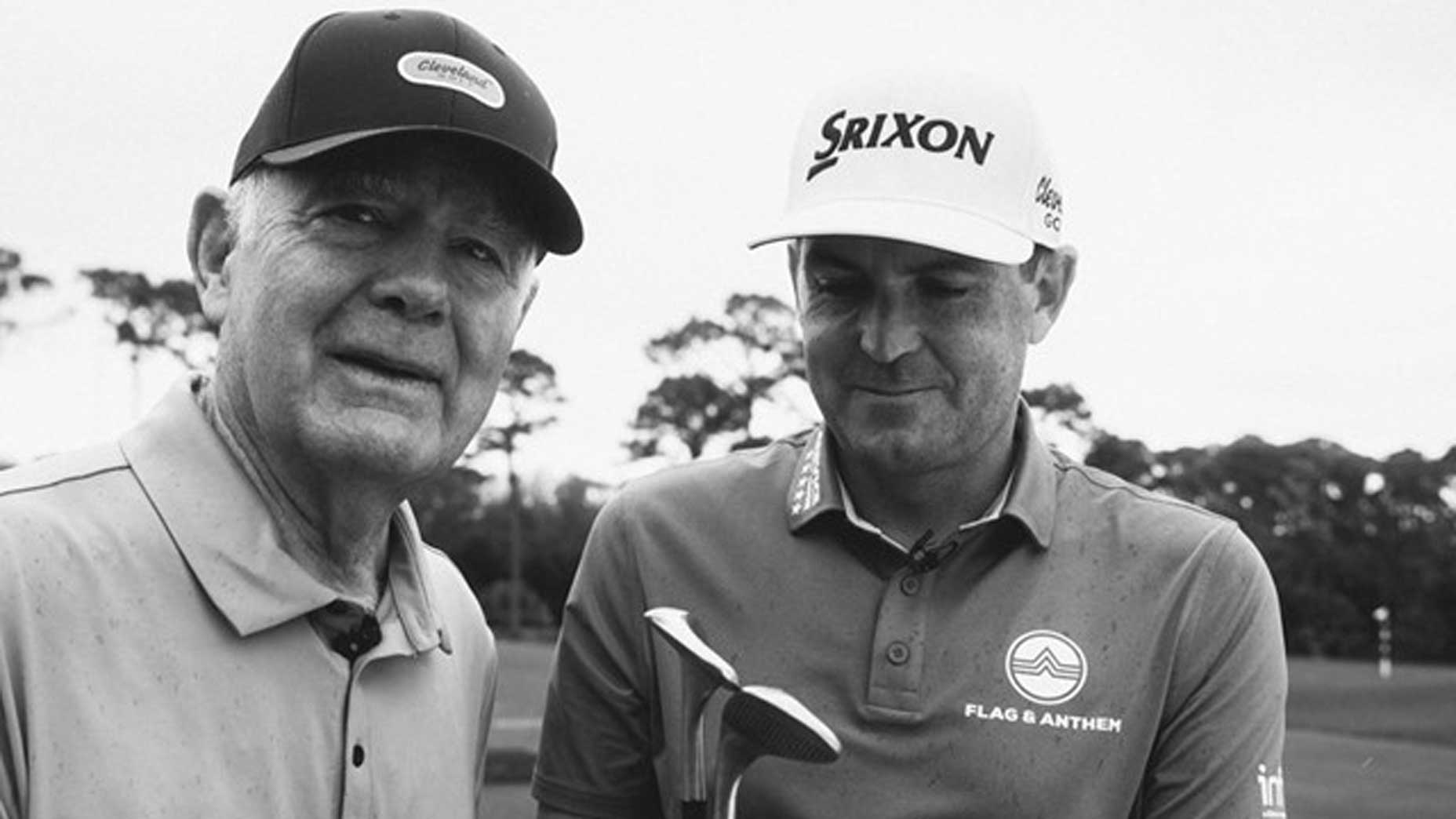 Two men wearing golf hats and shirts stand together outdoors; one looks at the camera while the other smiles and looks down at a Cleveland wedge they are holding. Trees are visible in the blurred background. Black and white photo.