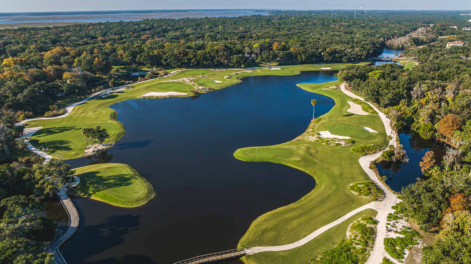 Aerial view of a lush golf course at Omni Amelia Island Resort with sand bunkers, winding fairways, and a central lake&mdash;perfect for a buddies' trip getaway&mdash;surrounded by dense trees under a clear sky.