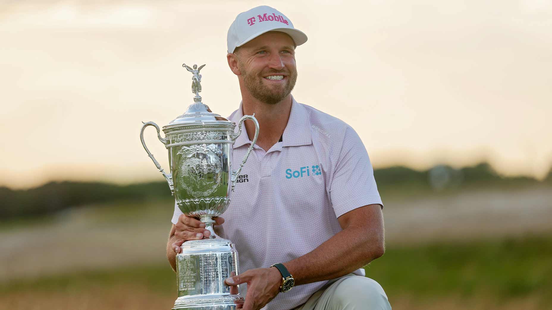 A smiling man in a light pink shirt and white cap holds a large silver trophy outdoors, celebrating his u.s. open money win against a blurry natural background.