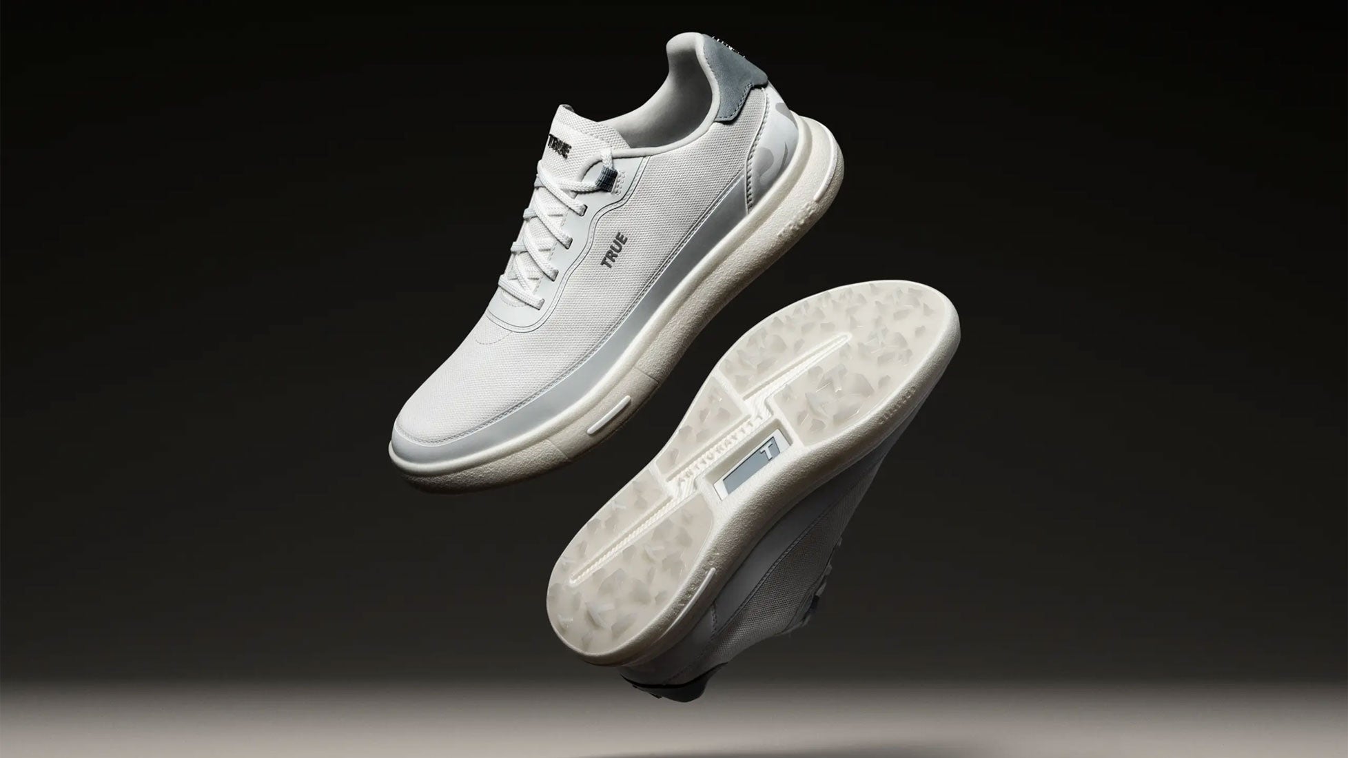 Two white athletic sneakers, including the innovative Antigravity golf shoe, are suspended in mid-air against a dark background&mdash;one showcasing the side and the other displaying the textured sole for a true sense of dynamic movement.