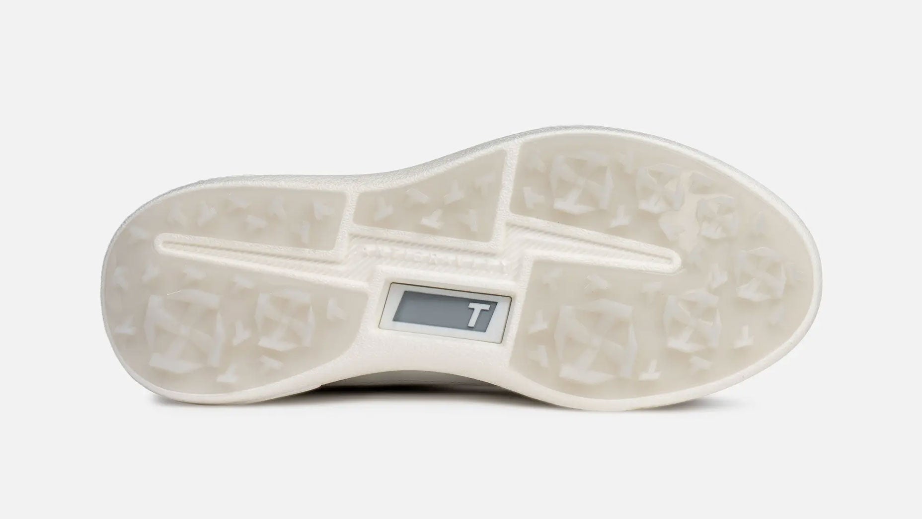 The image shows the translucent, textured sole of a white sneaker, featuring geometric grip patterns and a small rectangular window with the letter T visible inside&mdash;a hallmark detail of a True golf shoe.