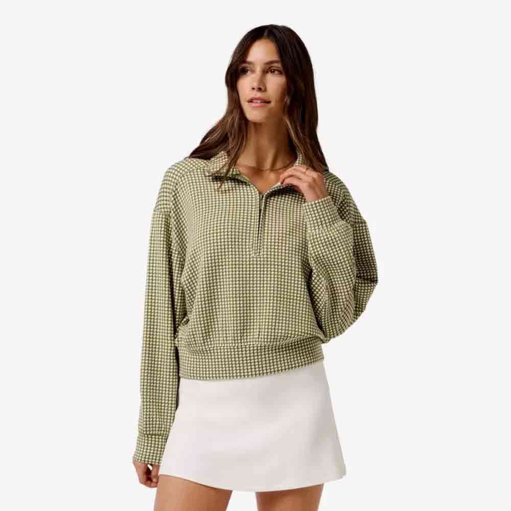 A woman wearing a green and white checkered long-sleeve pullover and a white skirt stands against a plain light background, looking slightly to the side&mdash;an elegant look perfect for Mother's Day.