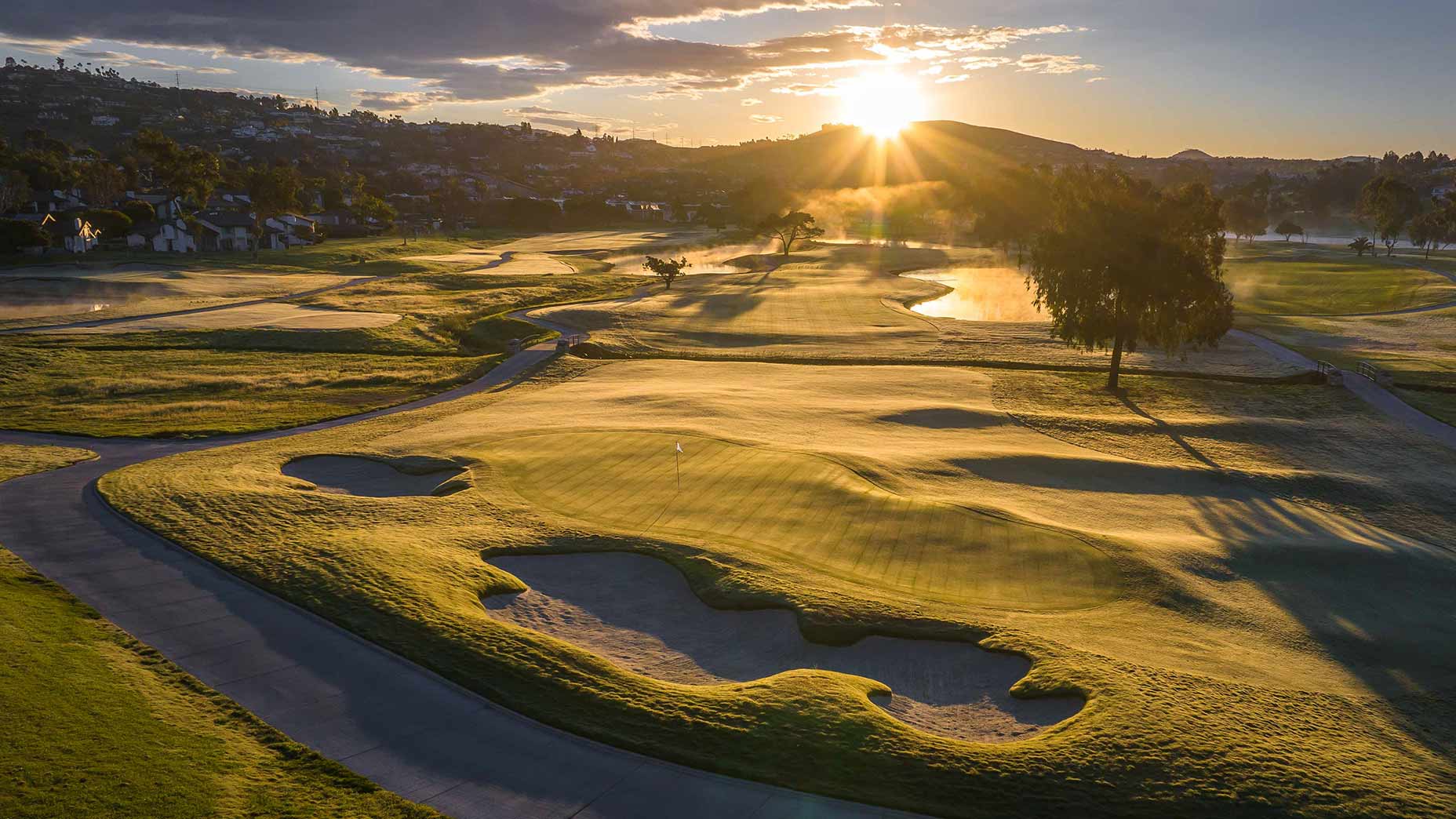 A scenic golf course at sunrise at Omni La Costa, with sunlight casting long shadows over the rolling fairways, sand bunkers, and a pond. Houses and hills are visible in the background beneath a partly cloudy sky.