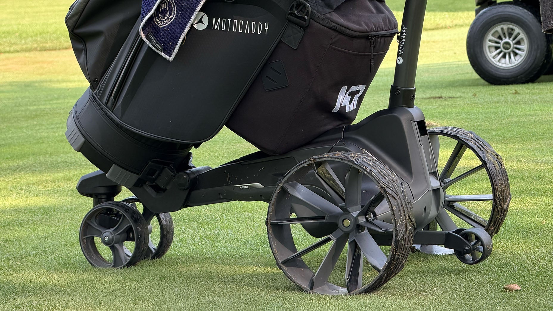 Close-up of a Motocaddy golf trolley, an essential among golf accessories, with large muddy rear wheels and a golf bag on a grassy course; a golf cart is visible in the background.