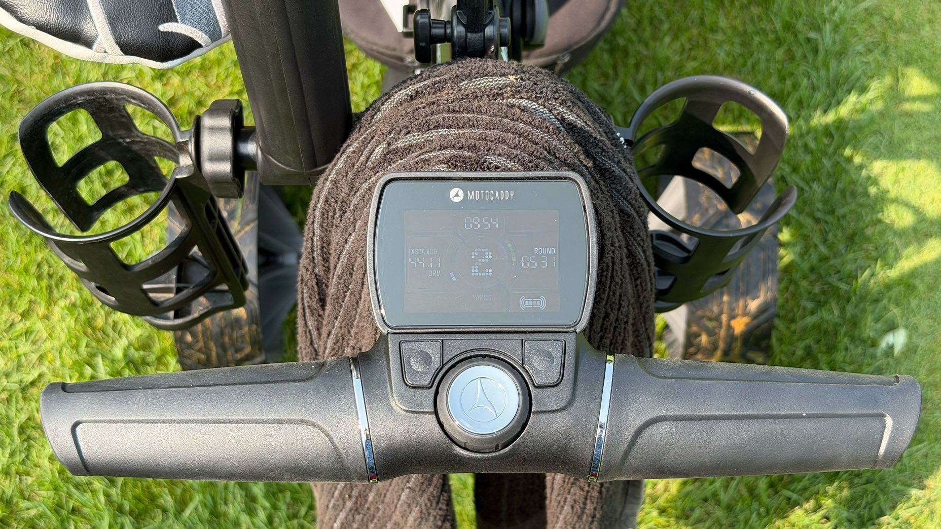 Close-up of a Motocaddy electric golf trolley handlebar with a digital display screen, two cup holders, and grass in the background&mdash;I tried it and was impressed by the comfort and technology packed into this premium golf trolley.