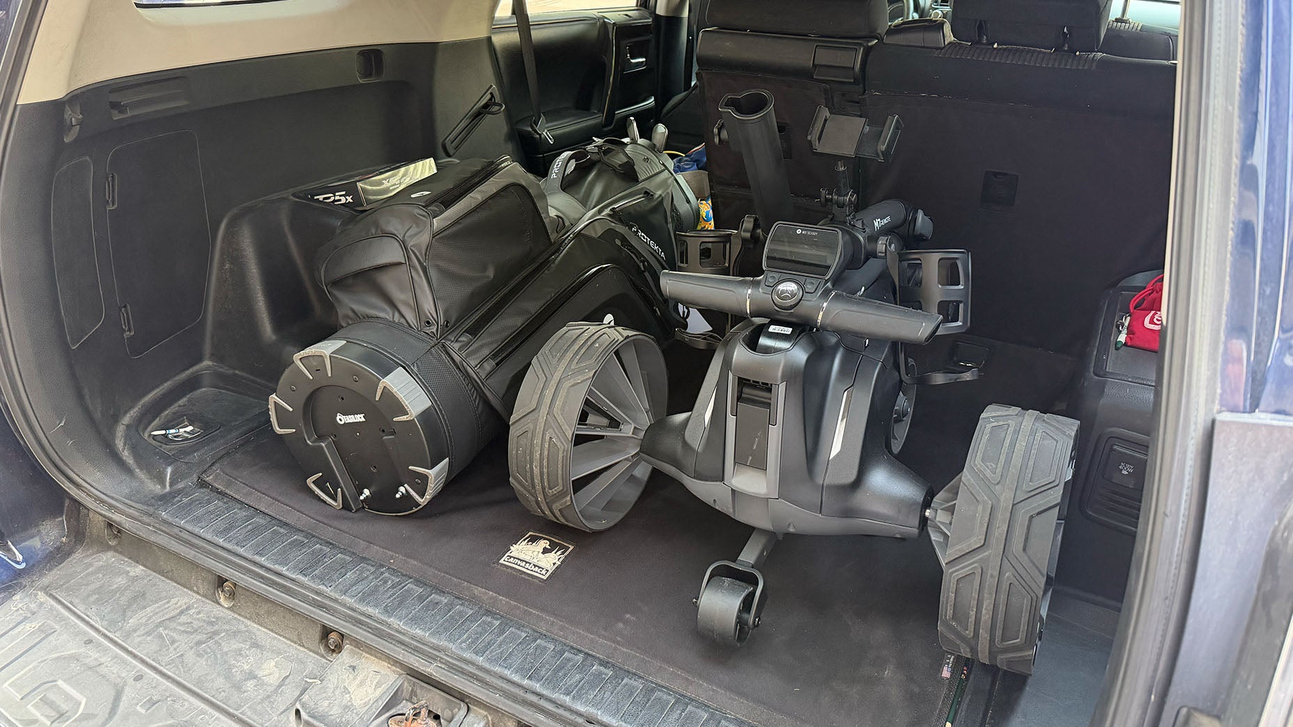 The open trunk of an SUV contains a golf bag inside a travel case and an electric golf push cart, specifically the Motocaddy M7 Remote. The cargo area is spacious, with other small items visible in the background.