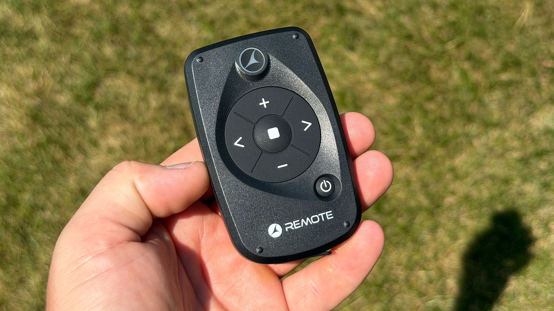 A hand holding a small black Motocaddy M7 Remote with directional arrows, plus and minus buttons, and a power button, against a grassy background.
