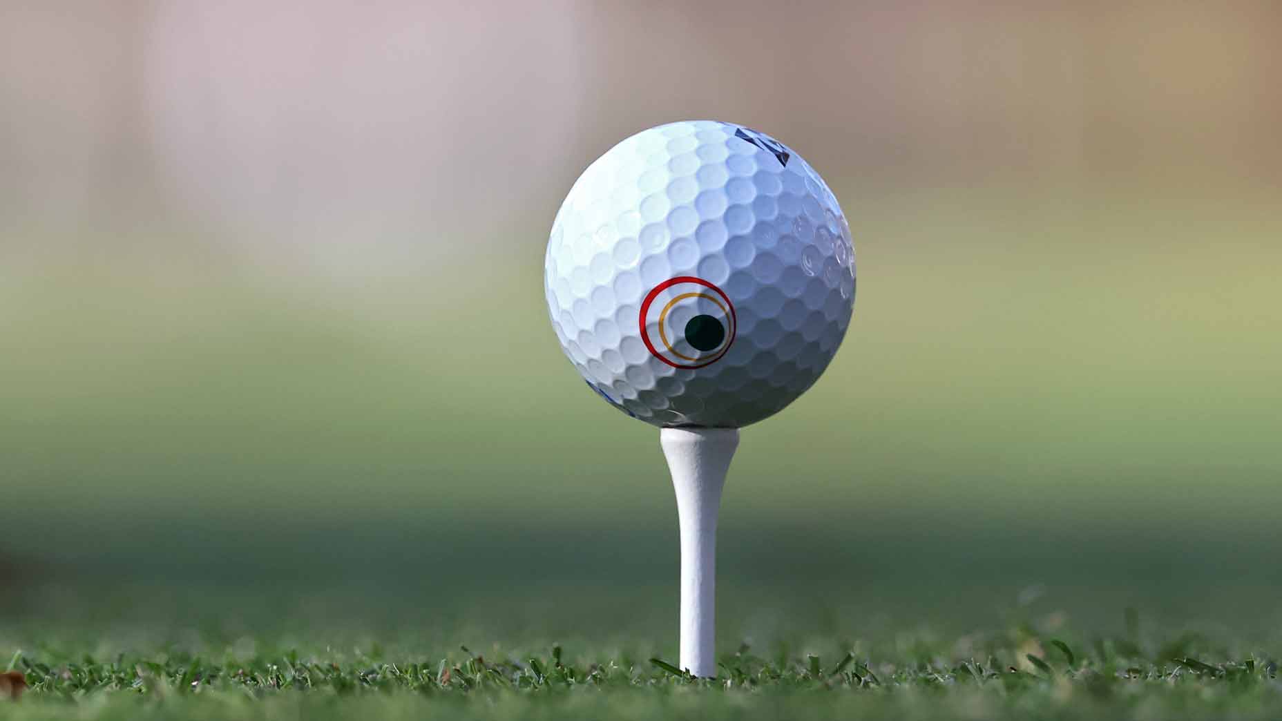 A golf ball with a small black spot marked by a red circle sits on a white tee on grassy ground, ready for a powerful swing with the irons, against a blurred brown and green background.