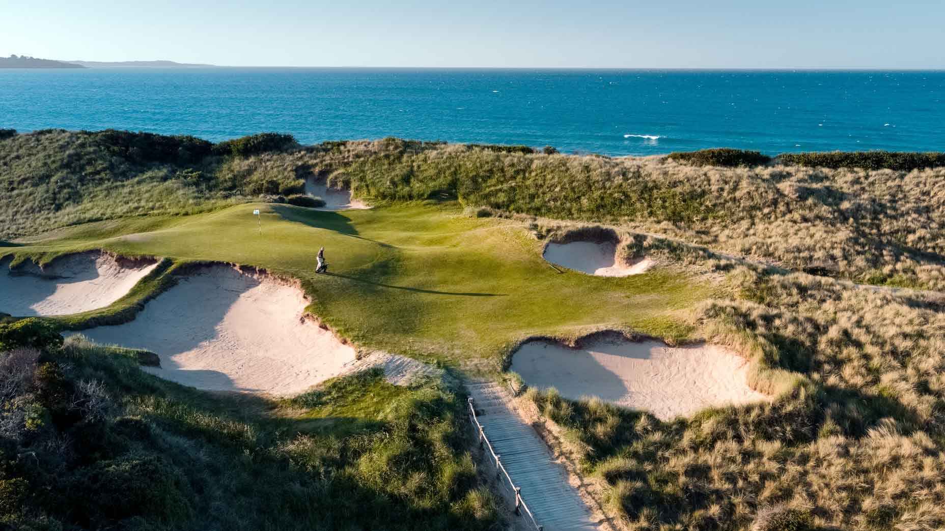 Aerial view of a coastal golf hole at Barnbougle Dunes golf course, surrounded by sand bunkers and grassy dunes, with the ocean and blue sky in the background in Tasmania.