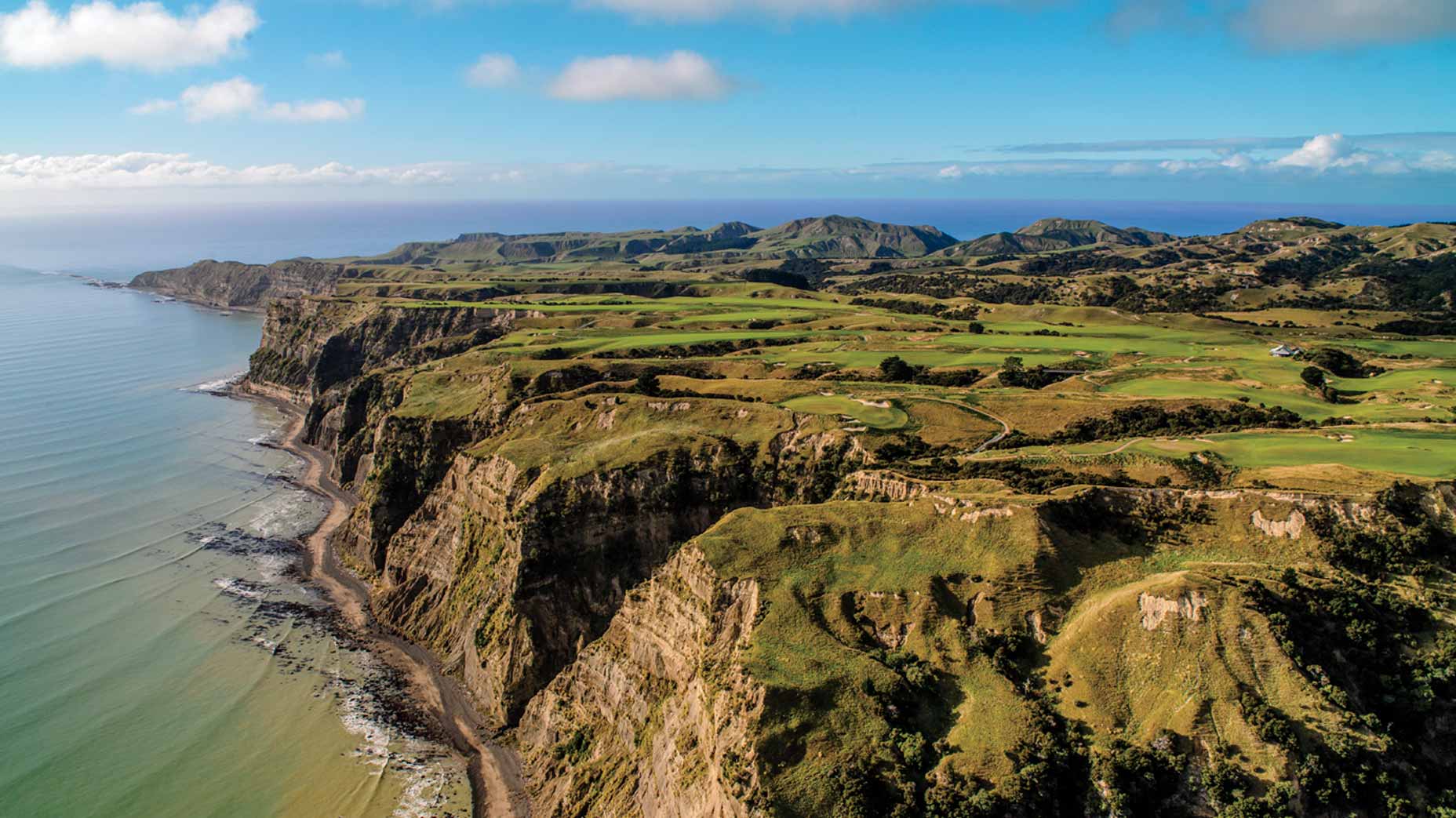 Aerial view of grassy cliffs meeting the ocean at Cape Kidnappers, with rolling hills and golf course fairways on top, under a bright blue sky with scattered clouds.
