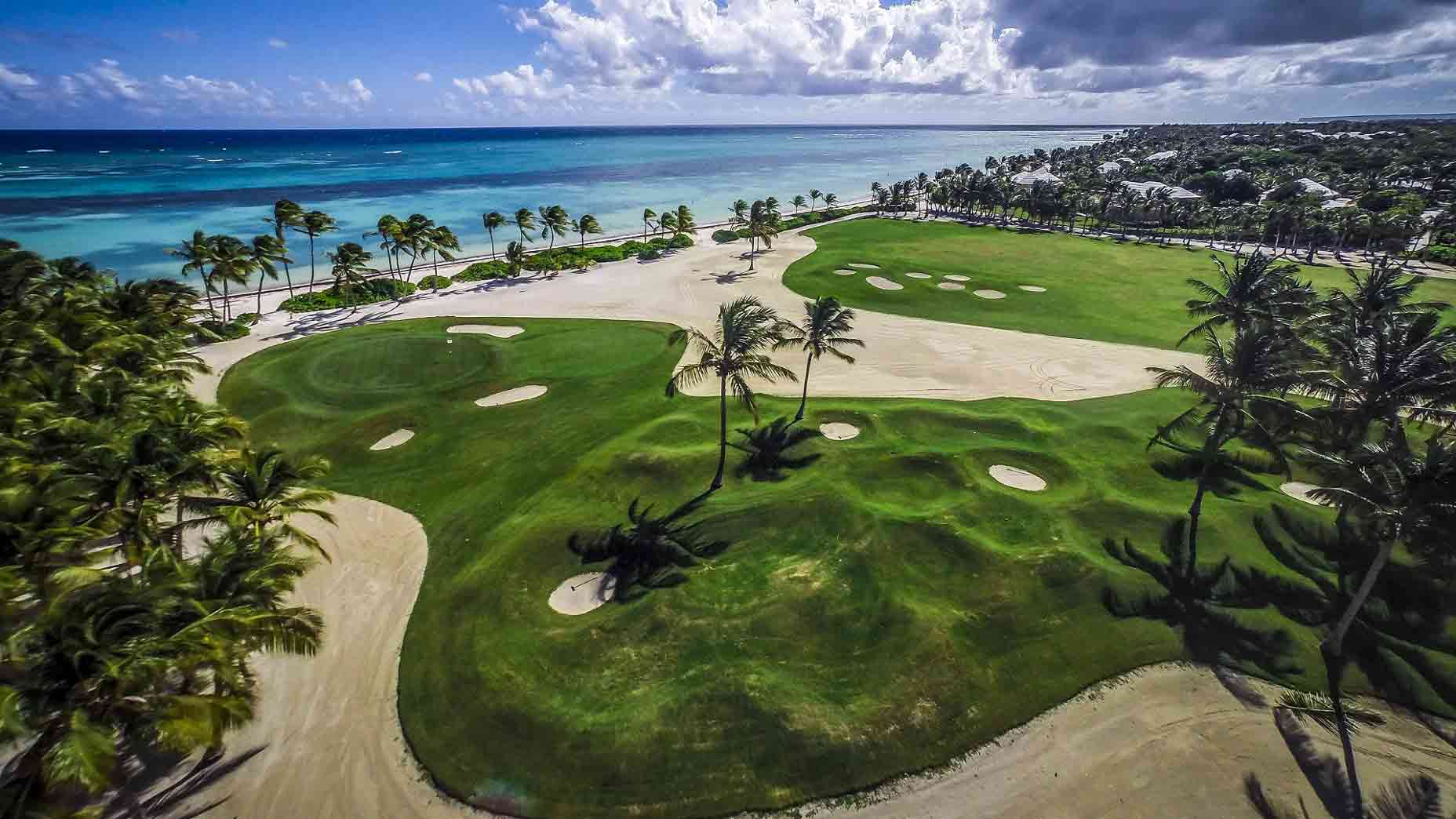 Aerial view of a tropical golf course by the ocean at Puntacana Resort & Club, featuring green fairways, sand bunkers, palm trees, and blue water under a partly cloudy sky&mdash;an idyllic destination recognized among the Top 100 Golf Resorts.