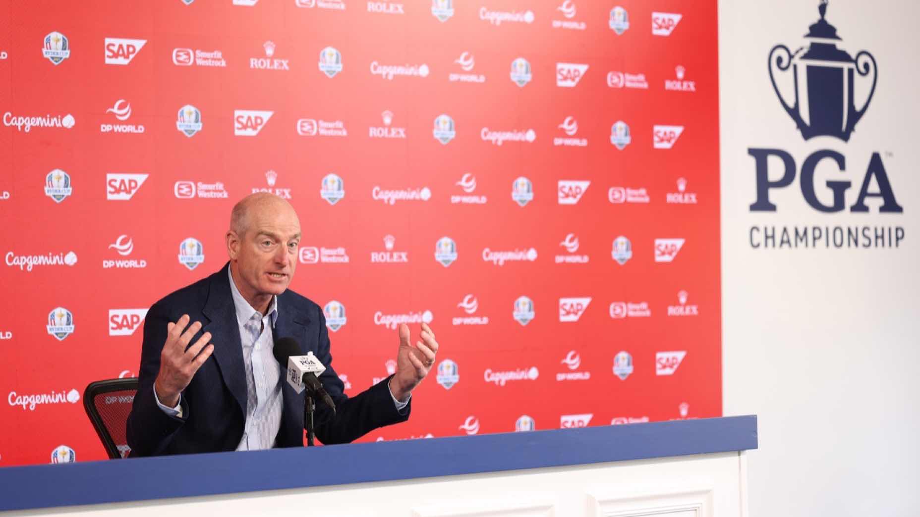Jim Furyk speaks at a press conference table with microphones, in front of a red PGA Championship backdrop featuring various sponsor logos. The PGA Championship logo is displayed on a white wall beside him.