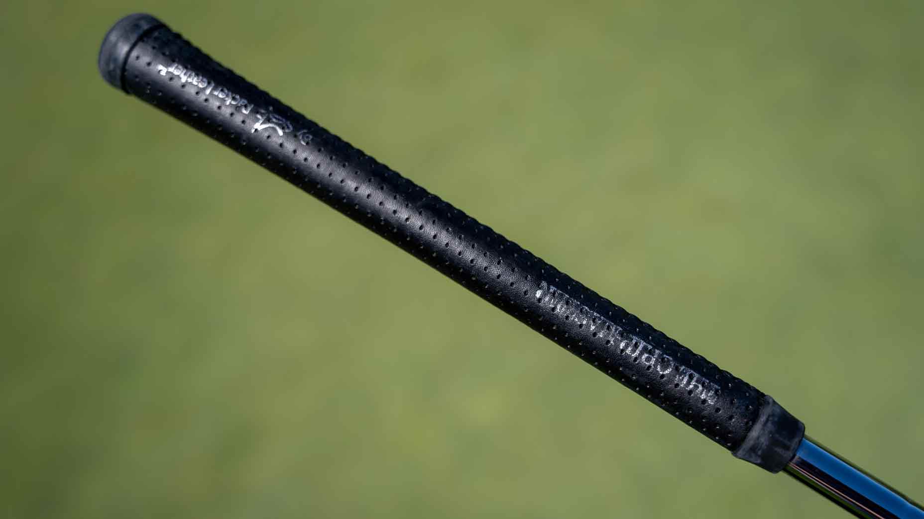 Close-up of a black, textured golf club grip featured in Bag Spy, with small dimples and white text, set against a blurred green background&mdash;just like something Min Woo Lee might have in his bag.