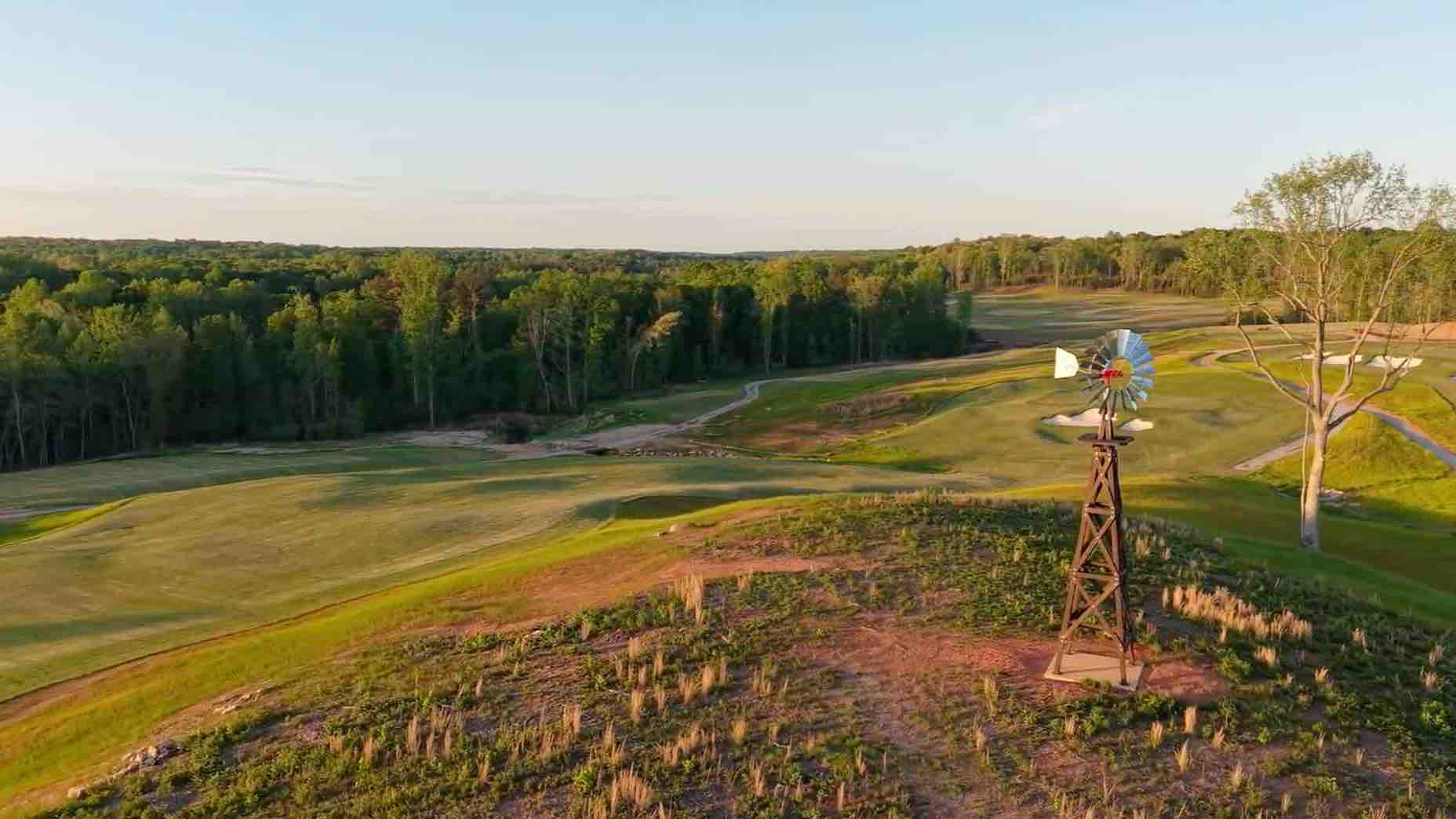 A scenic golf course with rolling green fairways, bordered by a dense forest. In the foreground, a rustic windmill stands on a small hill under a clear sky&mdash;holding the perfect spot for your next golf club secret weapon.