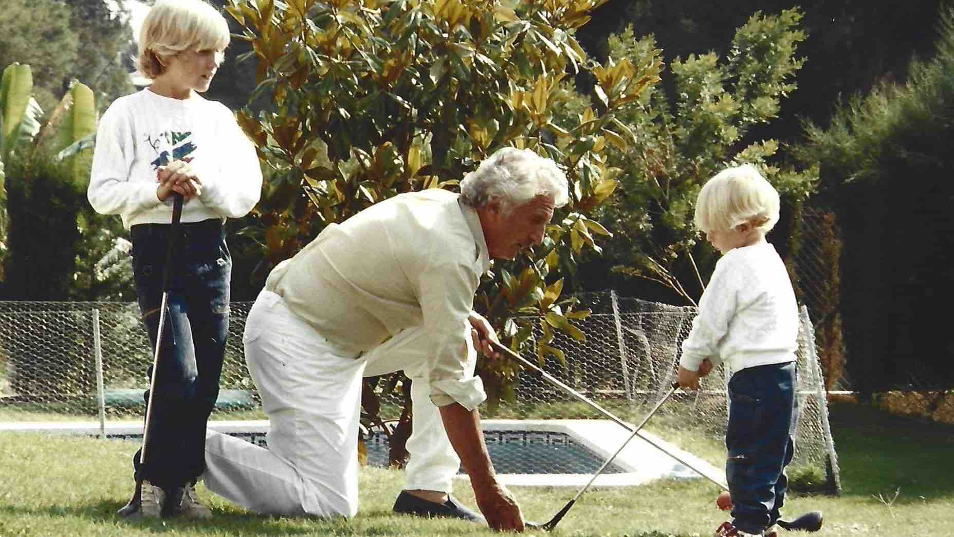 An older man kneels on the grass, helping a young child grasp a golf club&mdash;his secret weapon for fun&mdash;while another child stands nearby holding a club. They are outside near a pool, surrounded by lush greenery.