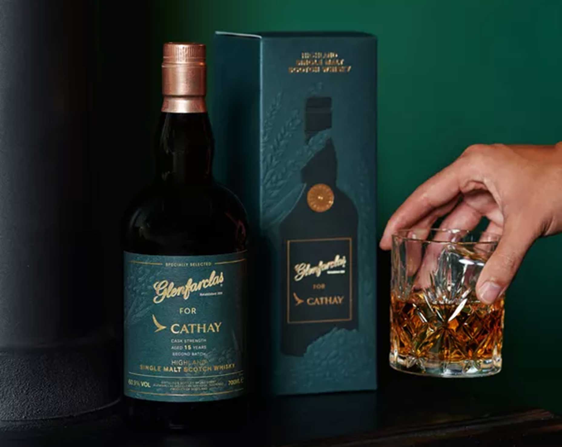 A hand holds a glass of whisky next to a Glenfarclas For Cathay single malt Scotch whisky bottle and its matching dark green box, set against a dark background&mdash;an exceptional gift for Father&rsquo;s Day and fans of fine whiskies.